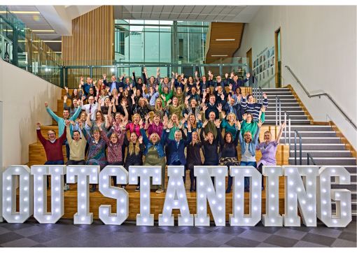 An image of five rows of students and lecturers, plus others in a lecture theatre, with a lit up Outstanding sign at the front