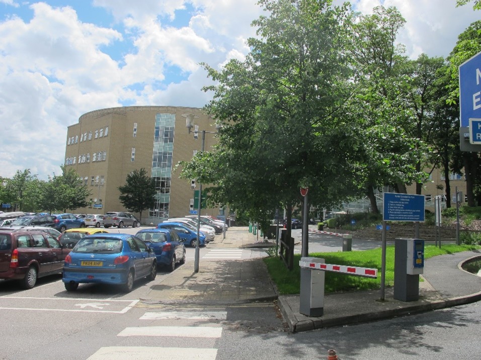 Cars are parked in front of the main hospital building. There are trees and a red and white parking barrier near a zebra crossing