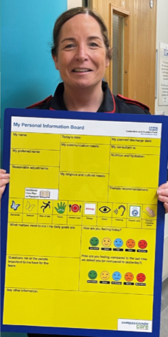 Photograph shows a smiling woman holding a bright yellow “My Parental Information Board” with a blue border, labelled sections, checkboxes, icons, and school branding. Board supports parent communication, featuring child-care information, routines, safety symbols, and coloured smiley-face feedback scales.