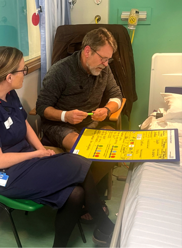 Photograph shows a hospital patient and healthcare worker reviewing a large yellow illustrated educational chart beside a hospital bed. Green clinical room, privacy curtain, seated adult wearing glasses, and chart’s colourful labels and diagrams support a patient-care discussion.