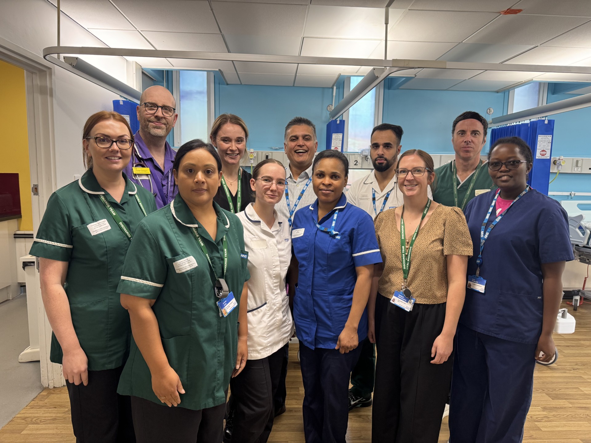Photograph shows a diverse group of healthcare professionals posing together in a hospital ward, wearing colourful scrubs, staff badges, and work attire. Blue privacy curtains, clinical equipment, ceiling lights, and hospital room surroundings provide context for a team or staff portrait.
