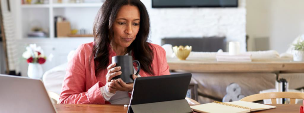 A woman sat at home in front of a tablet holding a mug with both hands.