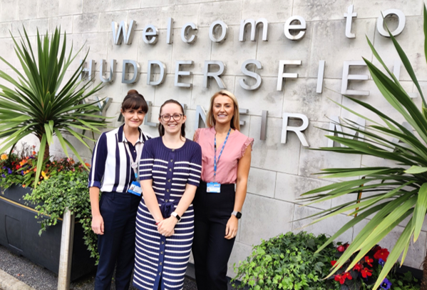 Three women standing in front of a concrete wall with large silver letters spelling "Welcome to Huddersfield Royal Infirmary." The scene includes two tall green plants and colourful flowers on either side, suggesting a welcoming or professional environment.