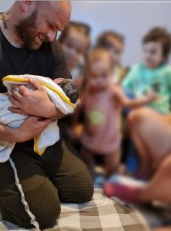 A father is kneeling on the floor, smiling at his newborn baby. Four children are in the background