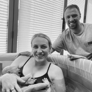 A happy woman in a birthing pool holding her new baby, with her partner behind her outside the pool.