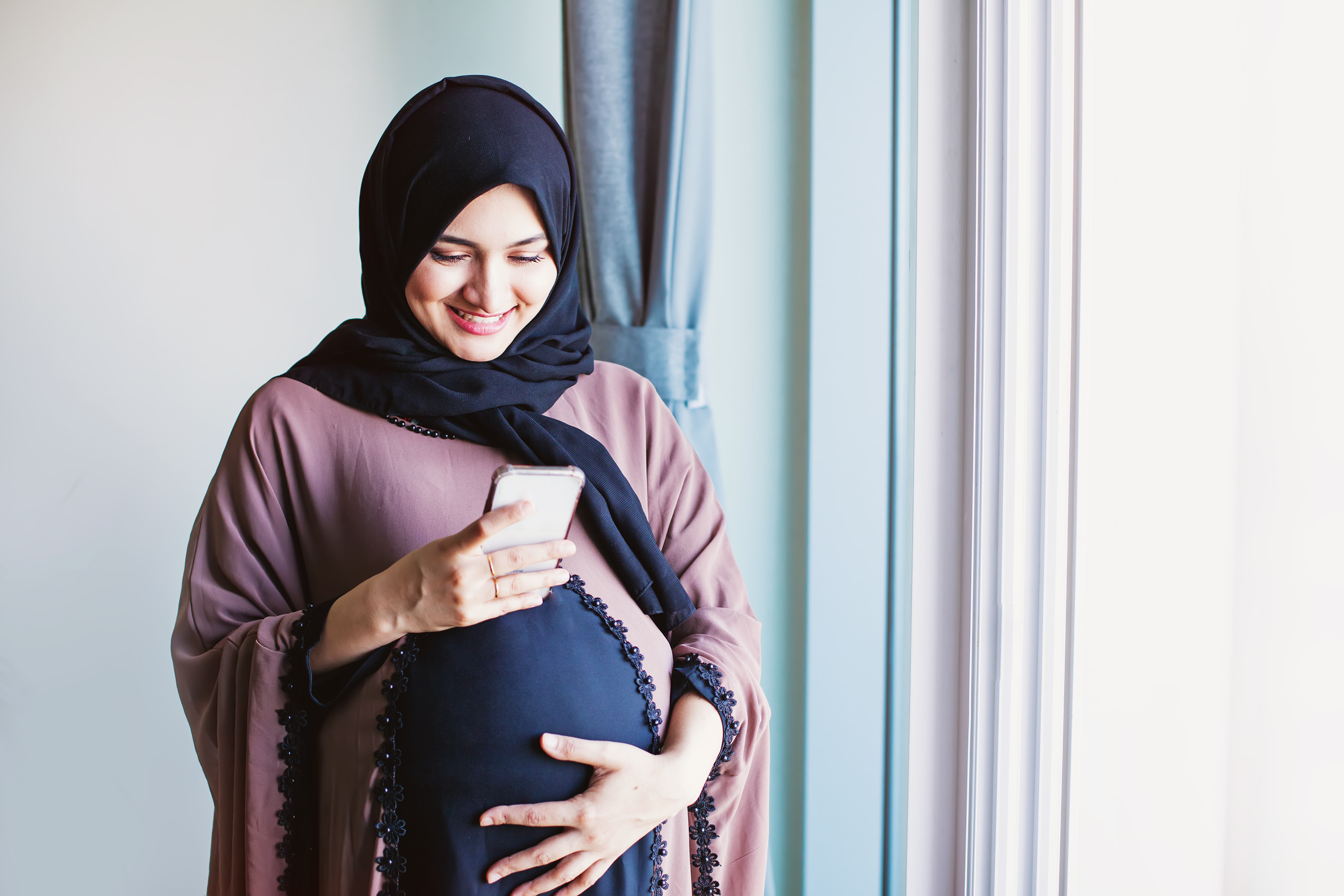 Pregnant woman wearing a black headscarf is standing by a window, using a smartphone and resting one hand on her baby bump.